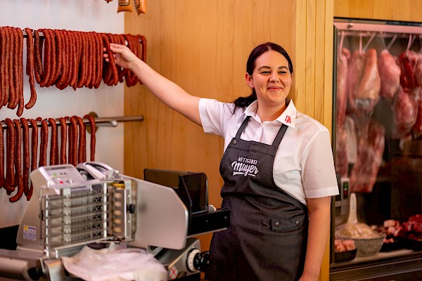 Das Bild zeigt eine Mitarbeiterin, die vor einer Schneidemaschine steht und ihre Hand nach einer darüberliegenden Wurst ausstreckt. Dabei lächelt sie in die Kamera