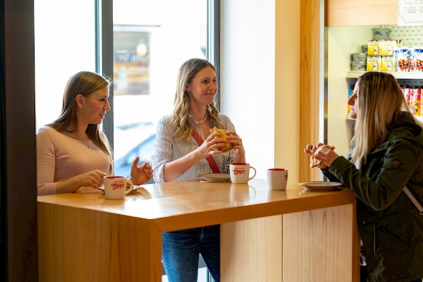 Drei Kundinnen stehen an einem Stehtisch und essen ein Fleischkäsebrötchen. Auch drei Kaffeetassen mit einem Schriftzug der Metzgerei Mayer stehen auf dem Tisch.
