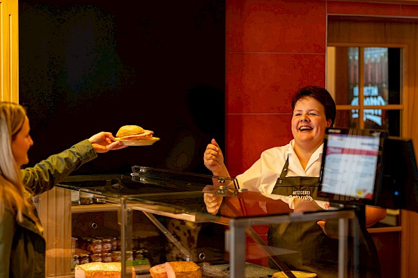 Eine Kundin nimmt ihr Fleischkäseweck auf einem Teller von einer lachenden Verkäuferin entgegen.