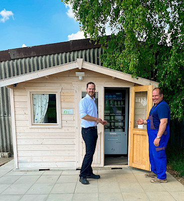 Tobias Fichtel und Bernd Breitenbücher stehen vor einem Hüttchen, in welchem sich ein Automat befinden.